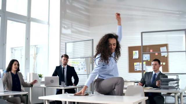 Young brunette businesswoman sweeping everything off from workplace in the office, standing on desk top and start dancing and throwing up papers in the air while colleagues joining her