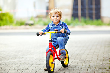 Active blond kid boy in colorful clothes driving balance and learner's bike or bicycle in domestic...