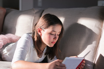 Hard light portrait of cute tween girl lying on sofa reading bookwith shadow on couch. High...