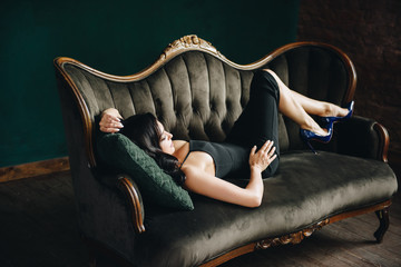 Beautiful girl in a black tight-fitting dress and heels. Black long wavy hair. Eastern appearance. Beautiful cut eyes. Brown sofa and green wall. A dark room. Sexy and stingy brunette. Portrait. - obrazy, fototapety, plakaty