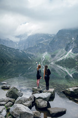 Two women, friends standing on the stony shore of lake Moskie Oko with scenic view of mountains with clouds and fog. Rysy mountains, Tatras. Poland, Slovakia. Rear view. Girl pointing on something © WellStock