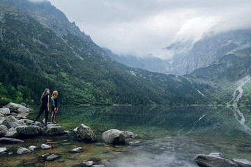 Two women, friends standing on the stony shore of lake Moskie Oko with scenic view of mountains with clouds and fog. Rysy mountains, Tatras. Poland, Slovakia. Rear view.