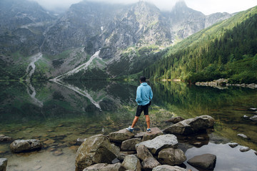 Young man in blue jacket standing on the stony shore of lake Moskie Oko with scenic view of mountains with clouds and fog. Rysy mountains, Tatras. Poland, Slovakia. Rear view © WellStock