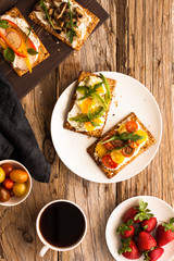 Breakfast table with tasty bruschetta crostini toasts, cream cheese, fresh vegetables and fruits and a cup of black coffee. Morning heathy balanced diet. Colorful ingredients on rustic wood background