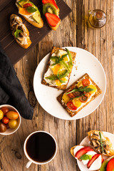 Breakfast table with tasty bruschetta crostini toasts, cream cheese, fresh vegetables and fruits and a cup of black coffee. Morning heathy balanced diet. Colorful ingredients on rustic wood background