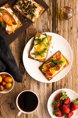 Breakfast table with tasty bruschetta crostini toasts, cream cheese, fresh vegetables and fruits and a cup of black coffee. Morning heathy balanced diet. Colorful ingredients on rustic wood background
