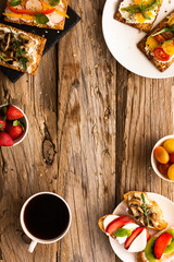 Breakfast table with tasty bruschetta crostini toasts, cream cheese, fresh vegetables and fruits and a cup of black coffee. Morning heathy balanced diet. Colorful ingredients on rustic wood background
