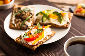 Breakfast table with tasty bruschetta crostini toasts, cream cheese, fresh vegetables and fruits and a cup of black coffee. Morning heathy balanced diet. Colorful ingredients on rustic wood background