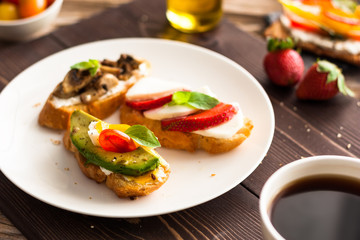 Fototapeta na wymiar Breakfast table with tasty bruschetta crostini toasts, cream cheese, fresh vegetables and fruits and a cup of black coffee. Morning heathy balanced diet. Colorful ingredients on rustic wood background