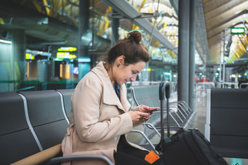 Business woman at the airport sitting waiting for her flight looking at the cell phone. Girl late for distressed plane flight at the international airport. Person sitting with suitcase on vacation sca