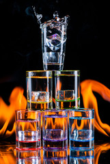 Multicolored glasses filled with alcoholic drinks, with splases of ice cubes falling inside, standing on the mirror surface on background of fire flames. On black. Conceptual design