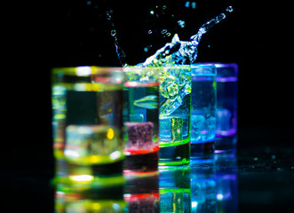 Multicolored glasses filled with alcoholic drinks, with splases of ice cubes falling inside, standing on the mirror surface. Black background. Conceptual, celebrated, commercial design