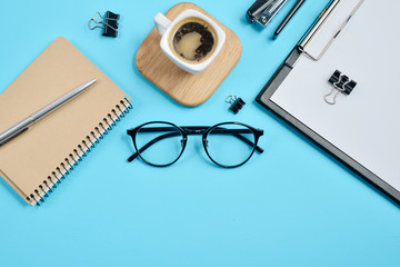 Office workspace with blank clip board, office supplies, pen, notepad, eyeglasses, stapler, coffee cup and spectacles on blue background. Flat lay, top view, stylish concept