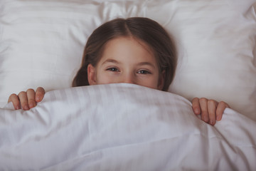 Obraz premium Close up of a cute happy little girl laughing, lying in her bed. Adorable little girl covering half of her face with a blanket. Cheerful kid having fun waking up in her bed