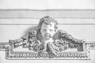 Fototapeta premium Closeup of an architectural detail. Bas-relief of a cherub with wings, enriched with stucco friezes, decorations, flowers and frames. Ancient sculpture attached to a plastered and painted wall.