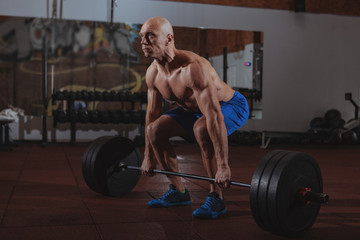 Fototapeta na wymiar Shirtless muscular male crossfit athlete lifting heavy barbell at the gym. Mature sportsman working out at crossfit box, lifting barbell