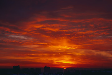 Cityscape with vivid fiery dawn. Amazing warm dramatic cloudy sky above dark silhouettes of city buildings. Orange sunlight. Atmospheric background of sunrise in overcast weather. Copy space.