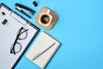 Office workspace with blank clip board, office supplies, pen, notepad, eyeglasses, stapler, coffee cup and spectacles on blue background. Flat lay, top view, stylish concept