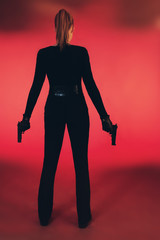 Girl with two guns dressed in black. Standing against red background.