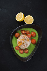 Bowl with baked hake medallions, green peas puree and cherry tomatoes. Flatlay on a black stone background, vertical shot