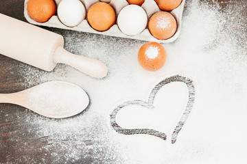Heart of flour on wooden desk, eggs, kitchenware