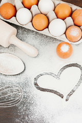 Heart of flour on wooden desk, eggs, kitchenware
