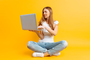 Portrait of a smiling young woman , sitting cross-legged using a laptop, on a yellow background