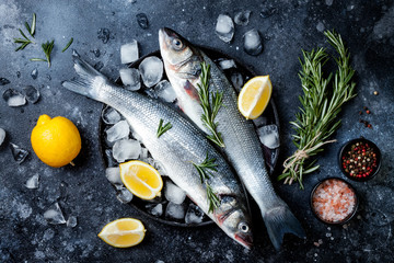 Fresh raw seabass fish on black stone background with spices, herbs, lemon and salt. Culinary seafood background with ingredients for cooking. Top view