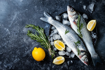 Fresh raw seabass fish on black stone background with spices, herbs, lemon. Culinary seafood background with ingredients for cooking. Top view