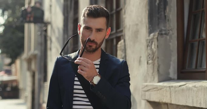 Portrait of the handsome Caucasian young man in stylish outfit taking off glasses and smiling to the camera at the street of nice old town.