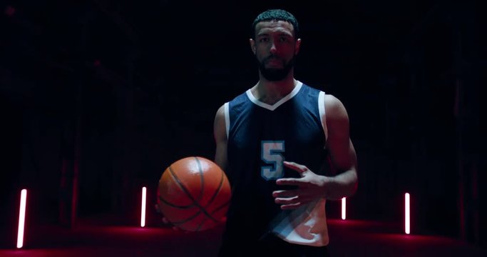 HANDHELD African American professional basketball player posing with a ball against dark background in a large abandoned warehouse. 4K UHD 60 FPS SLOW MOTION