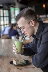 Young stylish man enjoys a cake and a drink while sitting in a cafe with large windows