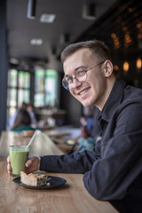 Young stylish man enjoys a cake and a drink while sitting in a cafe with large windows