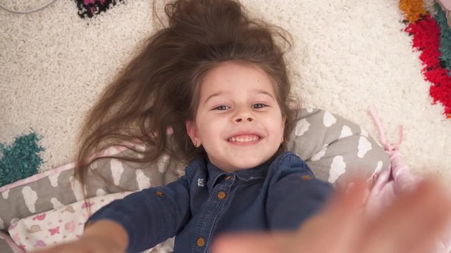 Top view, little girl lies on a soft white carpet and pulls her hands to the camera