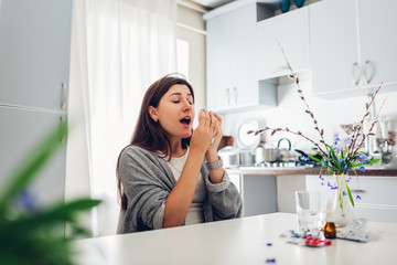 Spring allergy. Young woman sneezing because of flowers surrounded with pills on kitchen. Seasonal allergy concept. - obrazy, fototapety, plakaty