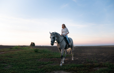 Young blonde girl riding on a horse on the field during sunset