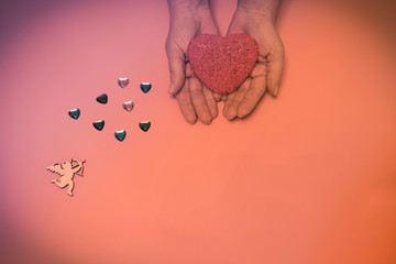 on a pink, beige background a red heart in human hands and an angel nearby.
