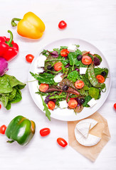 Salad in a white plate, cheese and vegetables on a white wooden background