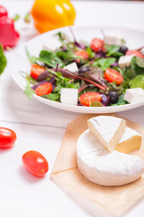 Salad in a white plate, cheese and vegetables on a white wooden background