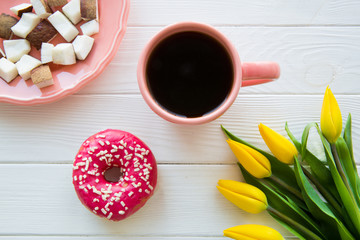 A cup of coffee, tasty doughnut and coconut pieces on white wooden table. Yellow tulip flowers, top view. Living coral color plate and cup. 