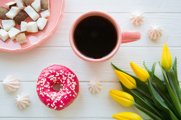 Obraz na płótnie Canvas Sweet breakfast. Pieces of coconut, morning black coffee and pink glazed doughnut. Yeelow tulip decoration on white wooden background. 
