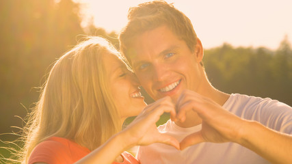 PORTRAIT: Happy man looks at camera while making a heart symbol with girlfriend