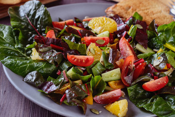 Salad with cutting leaves of chard, slices of tomatoes cherry, orange and cucumber in a grey plate with toast on a wooden table