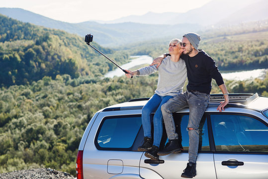 Young hipster couple in love making selfie using smart phone while sitting on the roof of off-road car on mountain background.
