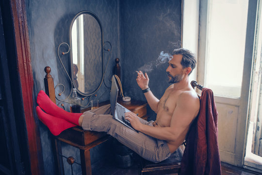 Freelance guy with a naked torso smokes and works on a laptop