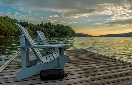 Two Light Blue Adirondack Chairs With A Black Boat Bumper Sits On A Floating Dock On Lake Massawippi At Sunrise Looking East