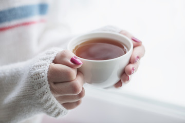 Tea drinking. The woman in a knitted, white pullover holds a cup of hot tea in her hands. Cozy weekends, winter drinks.