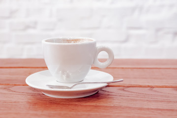 A white coffee cup with freshly brewed coffee and a spoon on a saucer stands on a dark wooden table near a white wall with a light shade on the table top in a cafe. Close up.