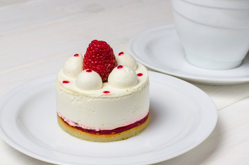 White airy cake with raspberry layer and raspberry berry lies on a white round plate next to a white cup, which stands on a white table
