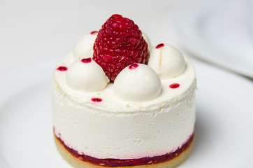 White airy cake with raspberry layer and raspberry berry lies on a white round plate next to a white cup, which stands on a white table